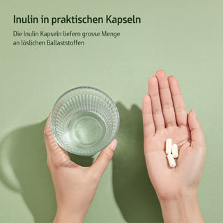 Zwei Hände halten Wasserglas und drei weiße Inulin-Kapseln vor grünem Hintergrund